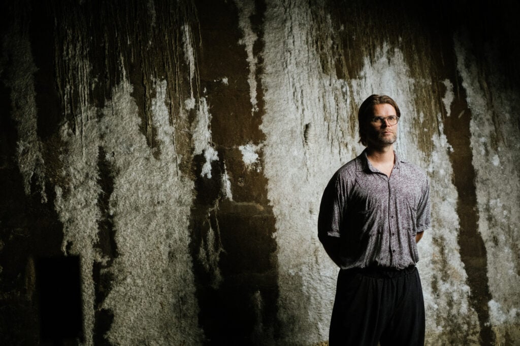 Jakob Kudsk Steensen standing against textured wall, dimly lit scene.