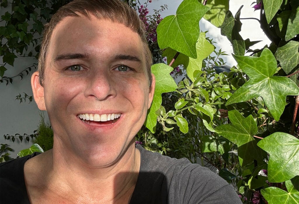 Photographer Kristian Kaarna in his own garden. Ivy leaves filter sunlight onto his face, creating a warm and relaxed atmosphere. The image reflects the humanity and positive spirit of the SmilingOulu26 project.