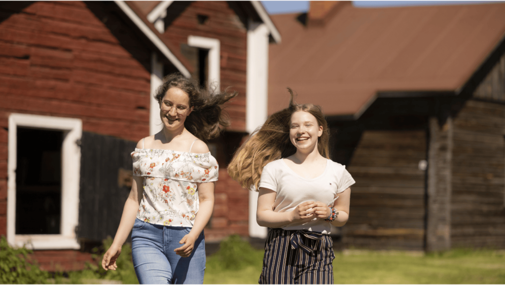 Kaksi naista kävelee aurinkoisella pihalla, punaiset talot.