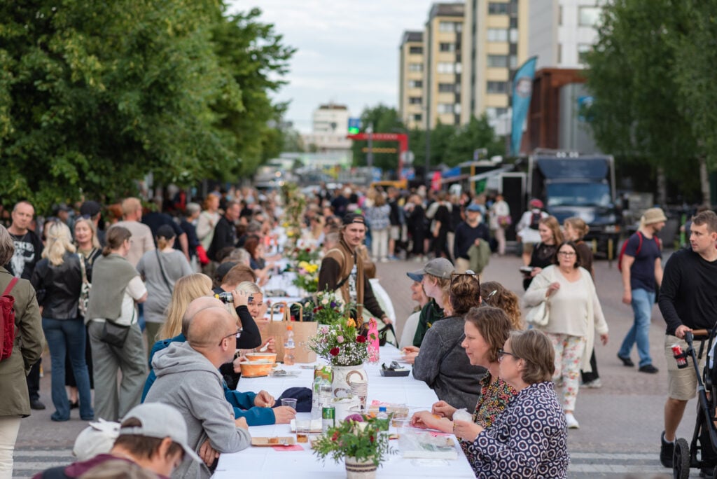 Ihmiset kokoontuneet pitkälle ruokapöydälle ulkoilmafestivaaleilla.