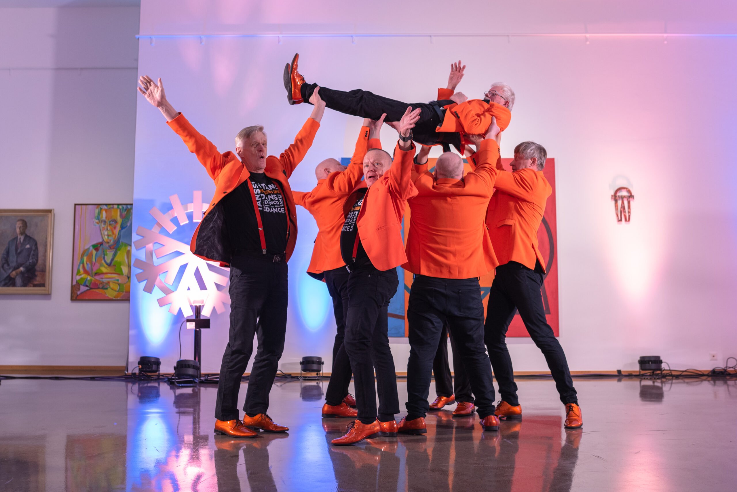 Miestanssijaryhmä Puumiehet Show tanssii oransseissa takeissa taidemuseossa.